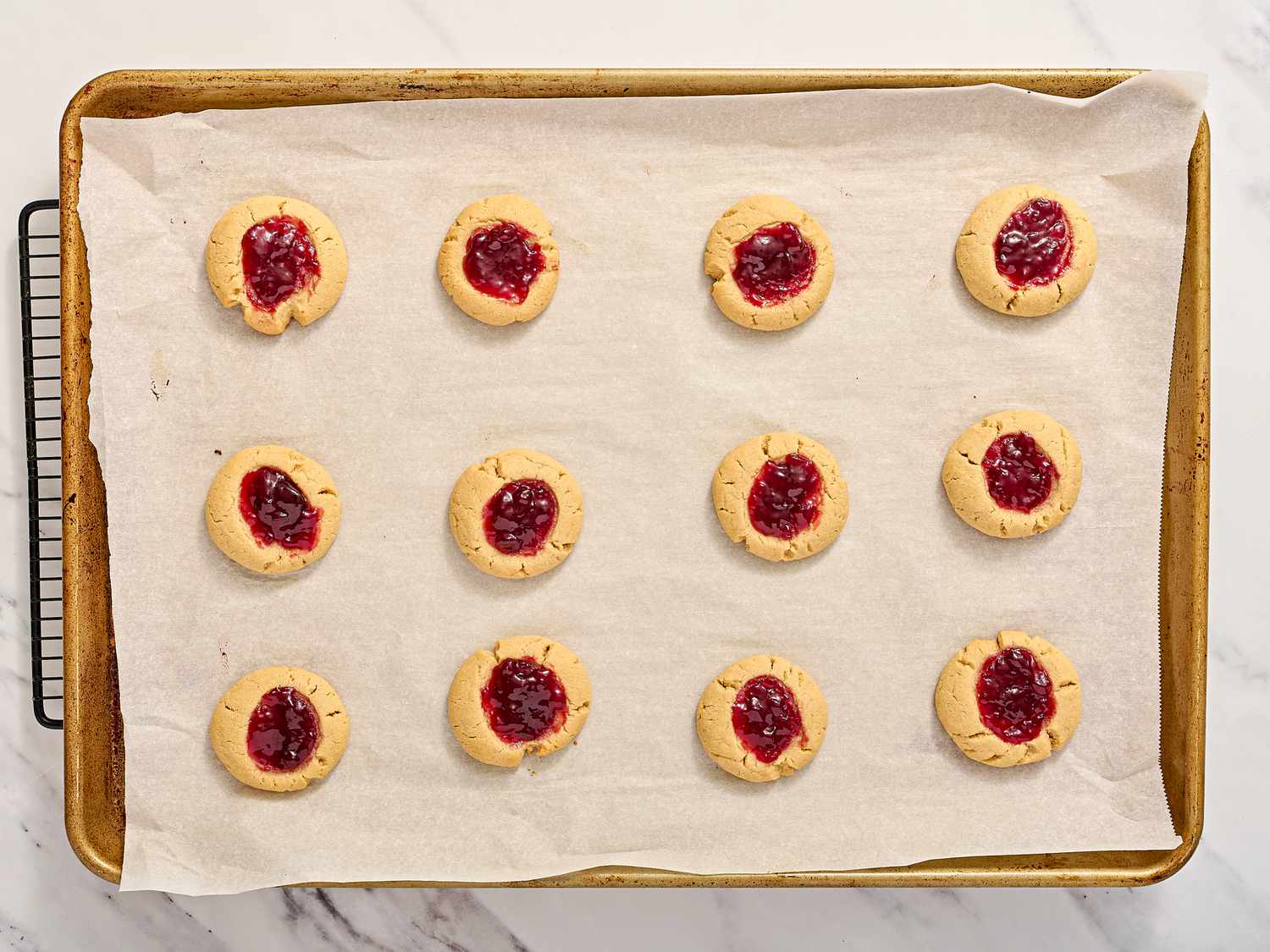 Baking tray with a dozen thumbprint cookies filled with jelly, arranged on parchment paper