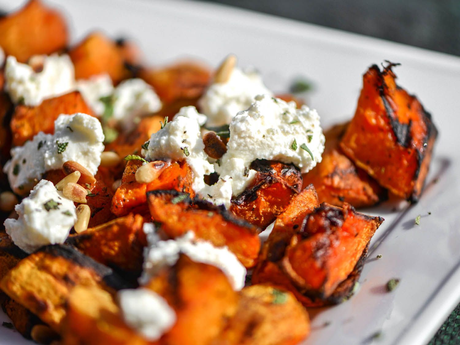 Close-up of grilled butternut squash cubes on a serving plate, garnished with ricotta and sage. 