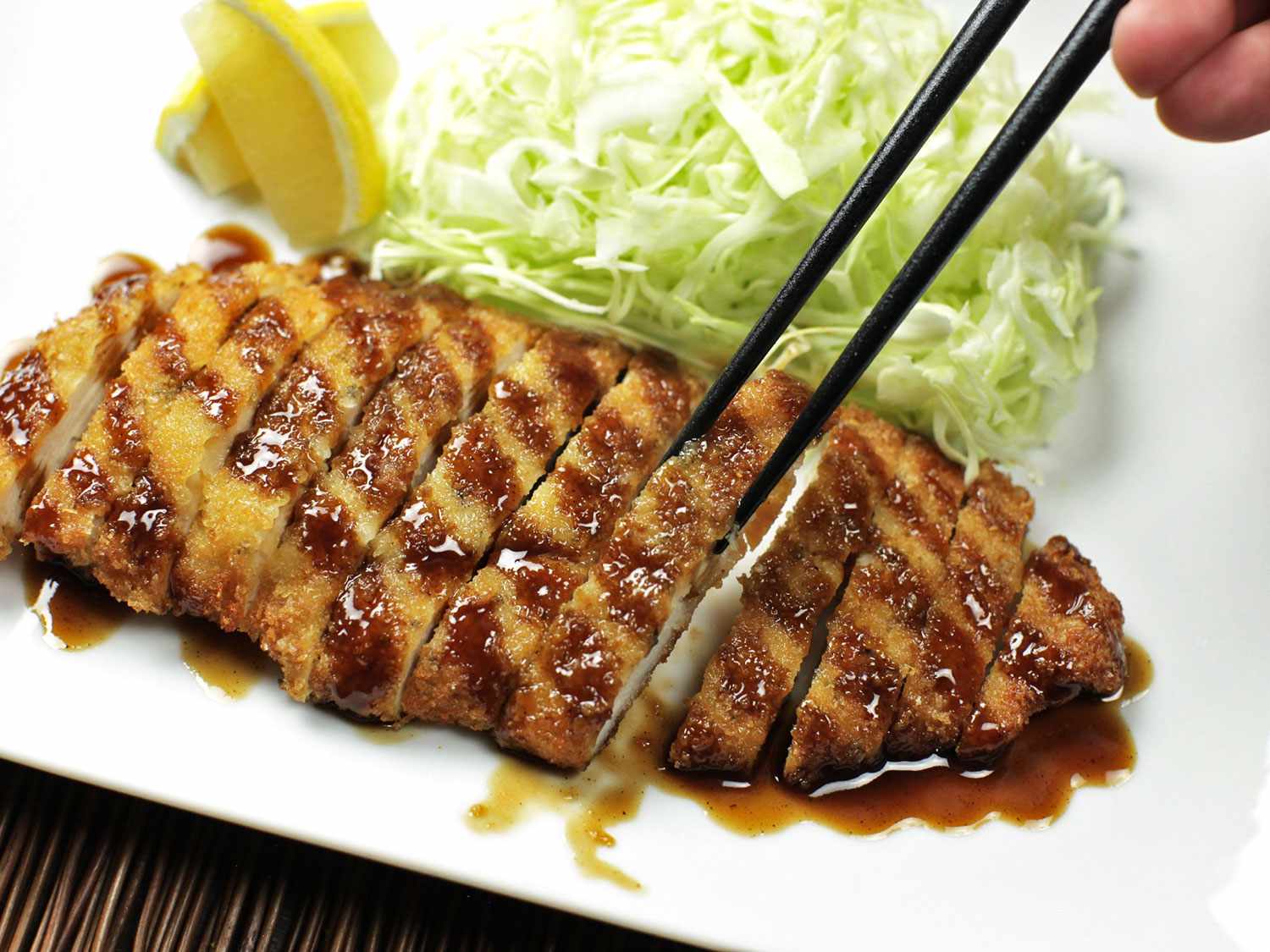 A sliced pork tonkatsu cutlet, served with shredded cabbage, lemon wedges, and a drizzle of tonkatsu sauce.