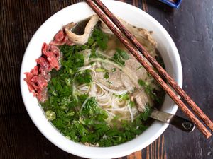 A bowl of pho with meat, noodles, herbs, and a pair of chopsticks resting on the side of the bowl