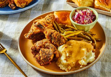 Dish of fried chicken with coleslaw, greenbeans, and mashed potatoes, with a fork and serving dishes in the background