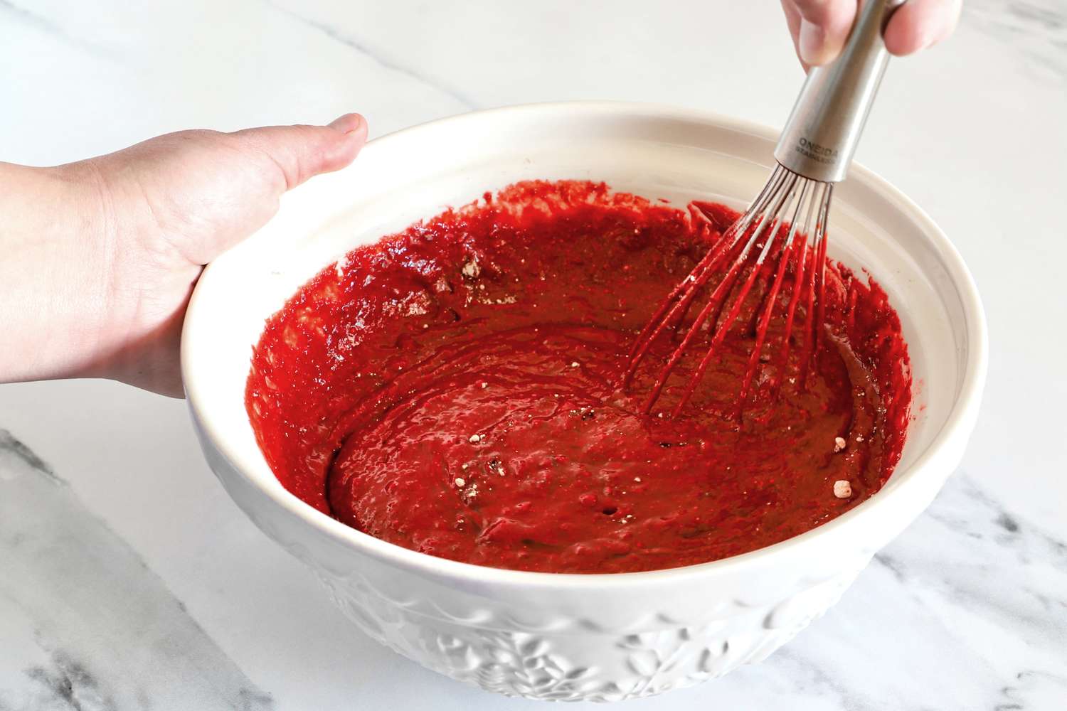 A hand stirring a red batter in a Mason Cash bowl with a wire whisk