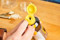 A hand holding and opening a bottle of liquid with a yellow cap, granules scattered on the countertop nearby
