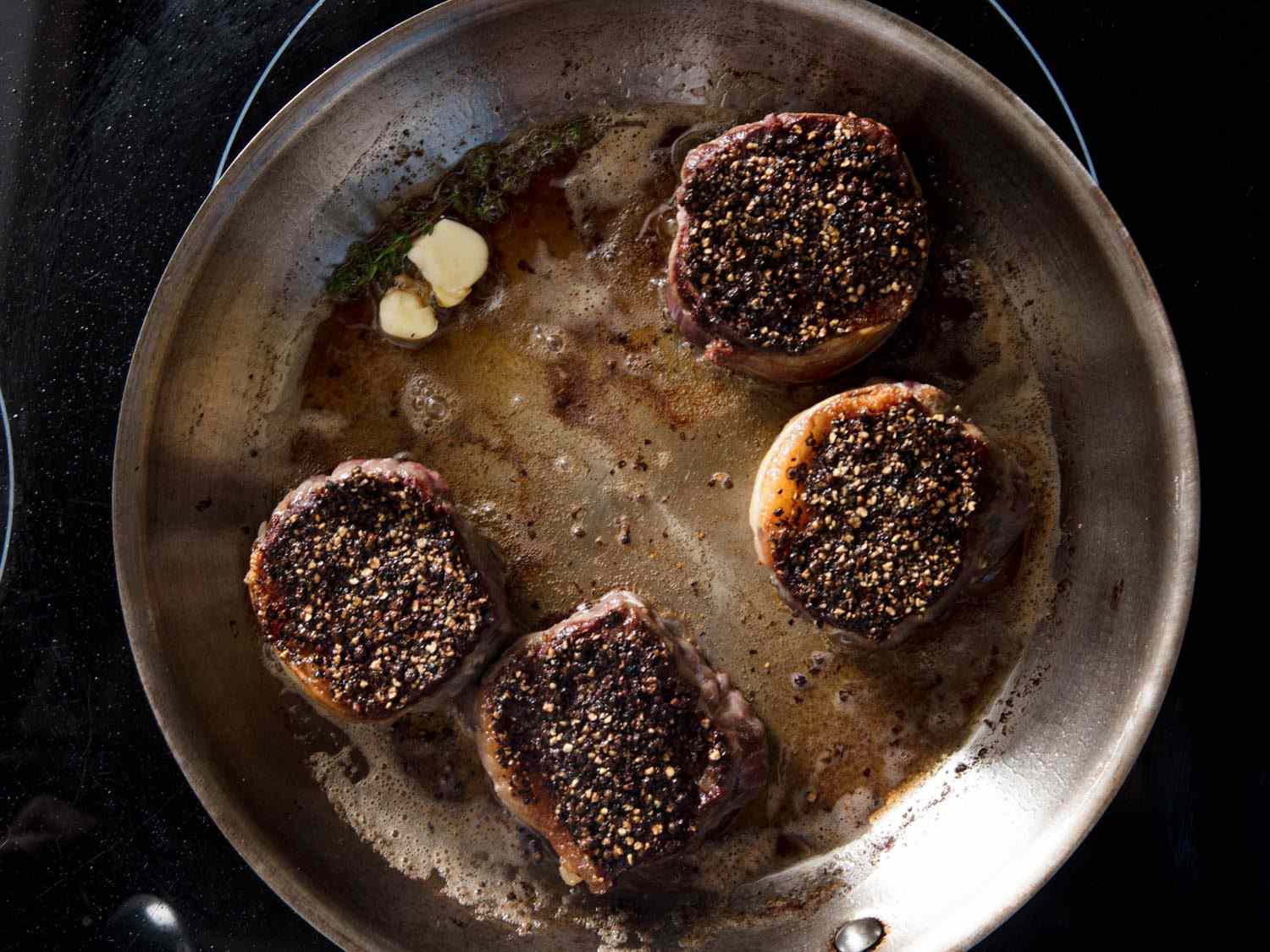 Overhead shot of pepper-crusted steaks being seared in a skillet. Butter and thyme have just been added to the pan.