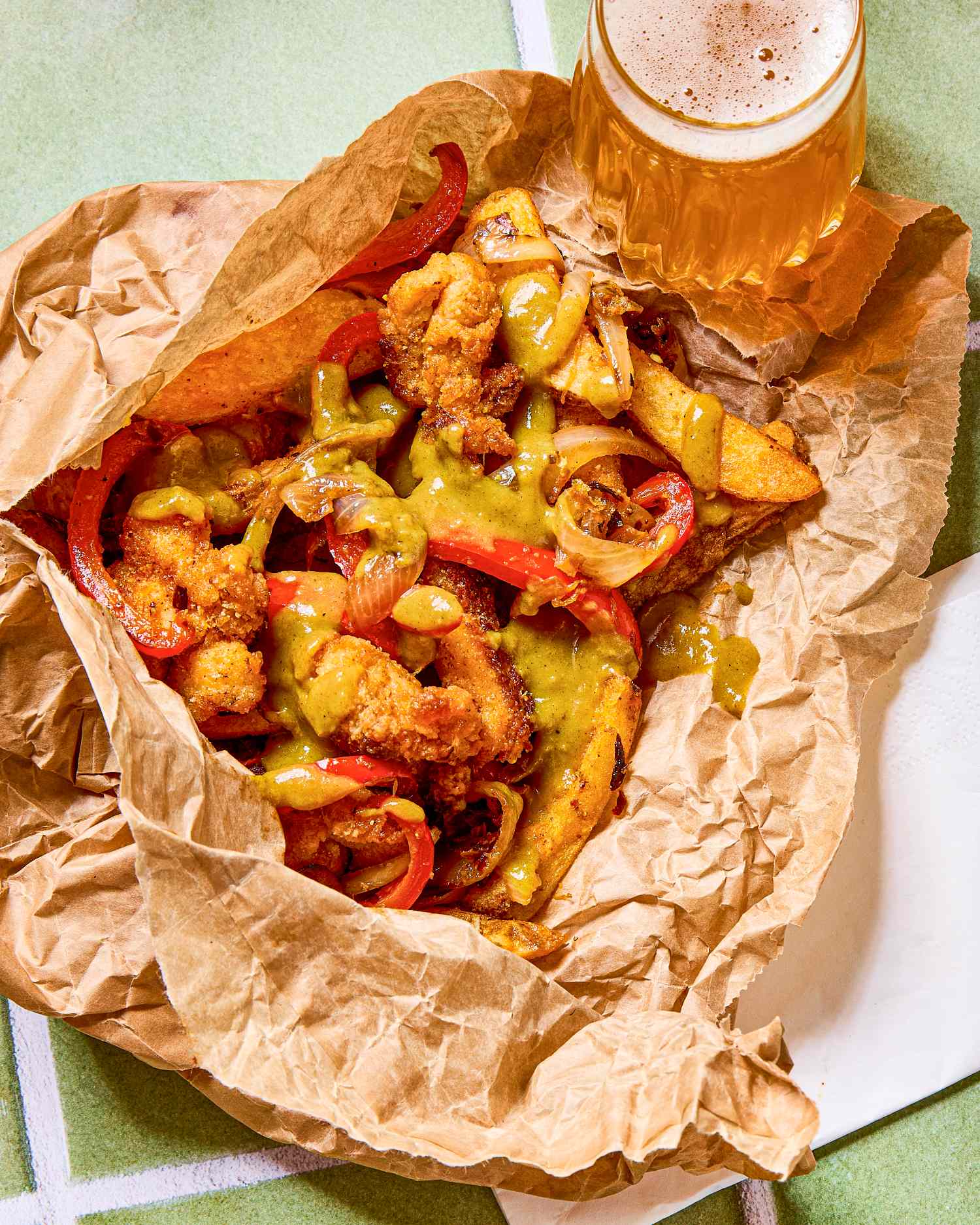 A serving of spice bag dish with fried chicken peppers fries and sauce in brown paper accompanied by a glass of beer on the side