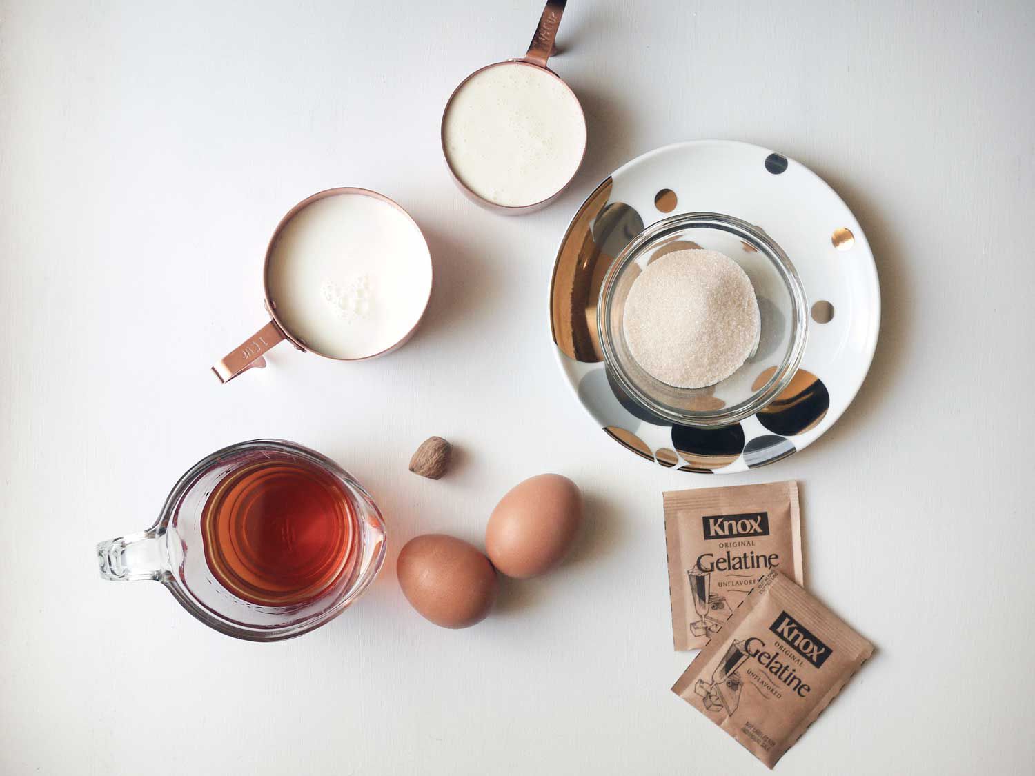 Overhead view of all the ingredients required to make eggnog jello shots, arranged on a white surface: rum, two eggs, nutmeg, 2 packs of gelatin, sugar, cream, milk. 