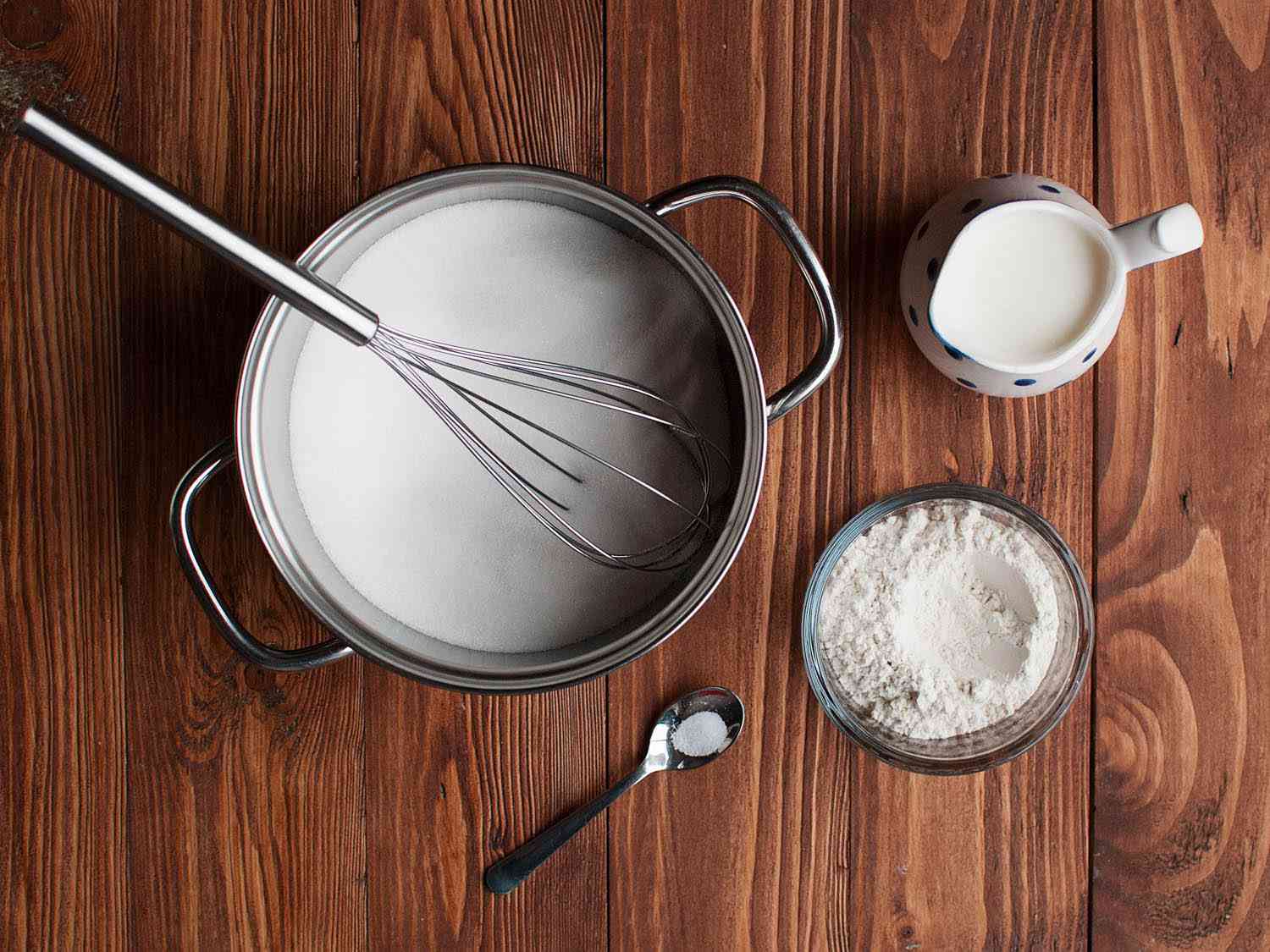 Ingredients for a pudding base gathered on a table: a pot containing milk and a whisk, a small spoon of salt, a bowl of flour and a jar of milk.