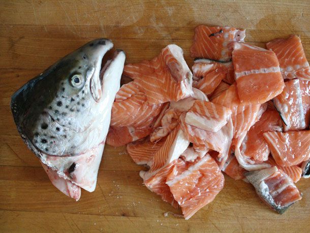 Overhead shot of a cutting board. A severed salmon head lies on the left side of the cutting board and assorted salmon scraps are piled on the right.