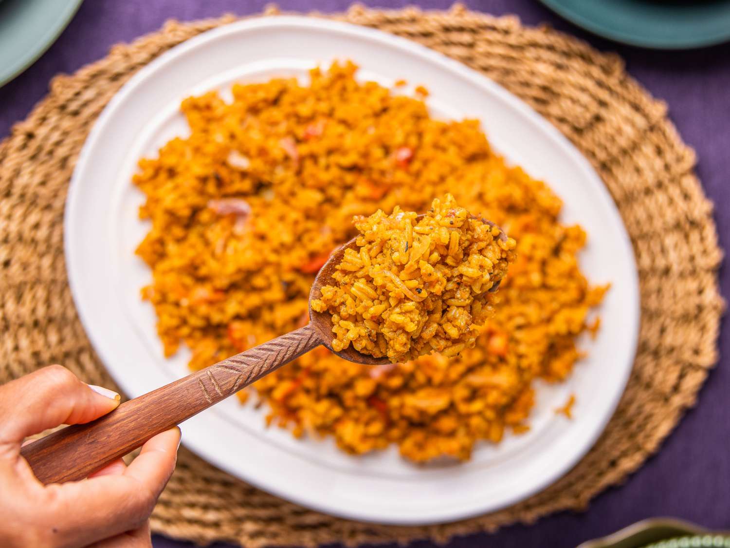 A plate of jollof rice with a wooden spoon holding a portion of rice placed on a woven placemat