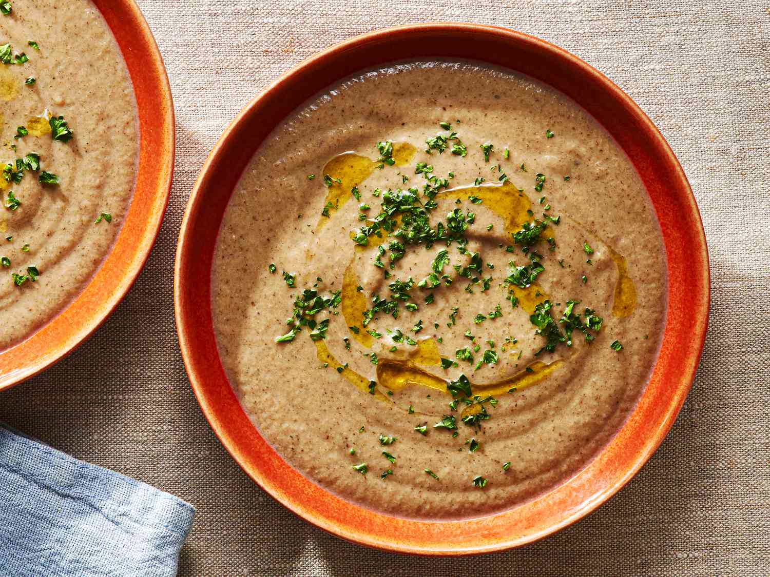 An orange-brown ceramic bowl holding creamy mushroom soup topped with chopped herbs and drizzled olive oil.