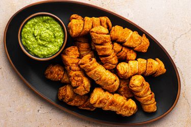 An oval black ceramic plate holding a pile of golden brown tequeÃ±os and a small bowl holding the avocado dipping sauce called guasacaca.