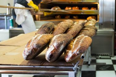 Loaves of bread on a baker's table in the kitchen at Tartine Bakery. In the background, someone is using a long handled peel to move loaves of bread in a commercial oven. 