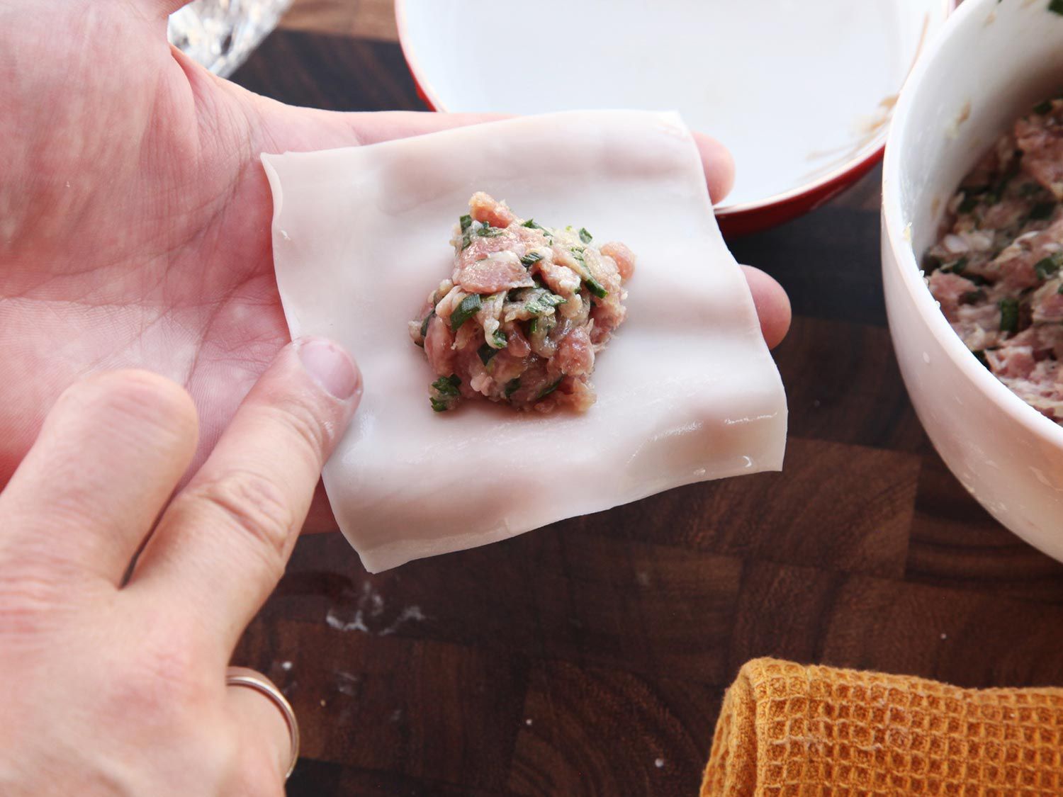 Moistening the edge of a wonton wrapper with a damp finger. There is a small amount of pork filling in the center of the wonton wrapper and a bowl of more filling nearby. 