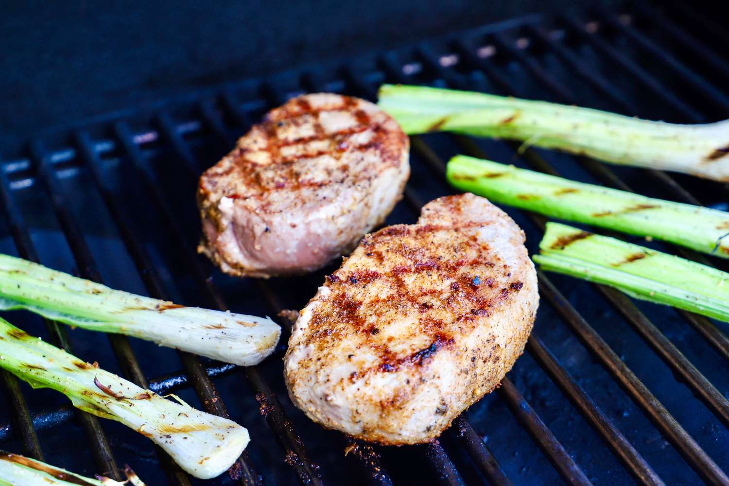 Two grilled Butcherbox meat cuts beside charred green onions on a barbecue grill