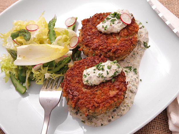 Two crab cakes topped with tartar sauce and sliced radish on a white plate with a side salad. 
