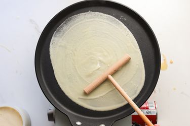 A person using a spreader to spread crepe batter onto a crepe pan.