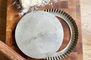 A metal tart pan on a wooden cutting board with pie dough and a rolling pin next to it