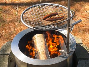 Hot dogs cooking on a grill over a flaming outdoor firepit