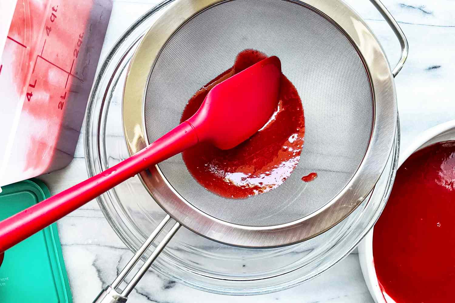 A red spatula pushing a thick liquid through Rösle Stainless Steel Round Handle Kitchen Strainer over a clear bowl