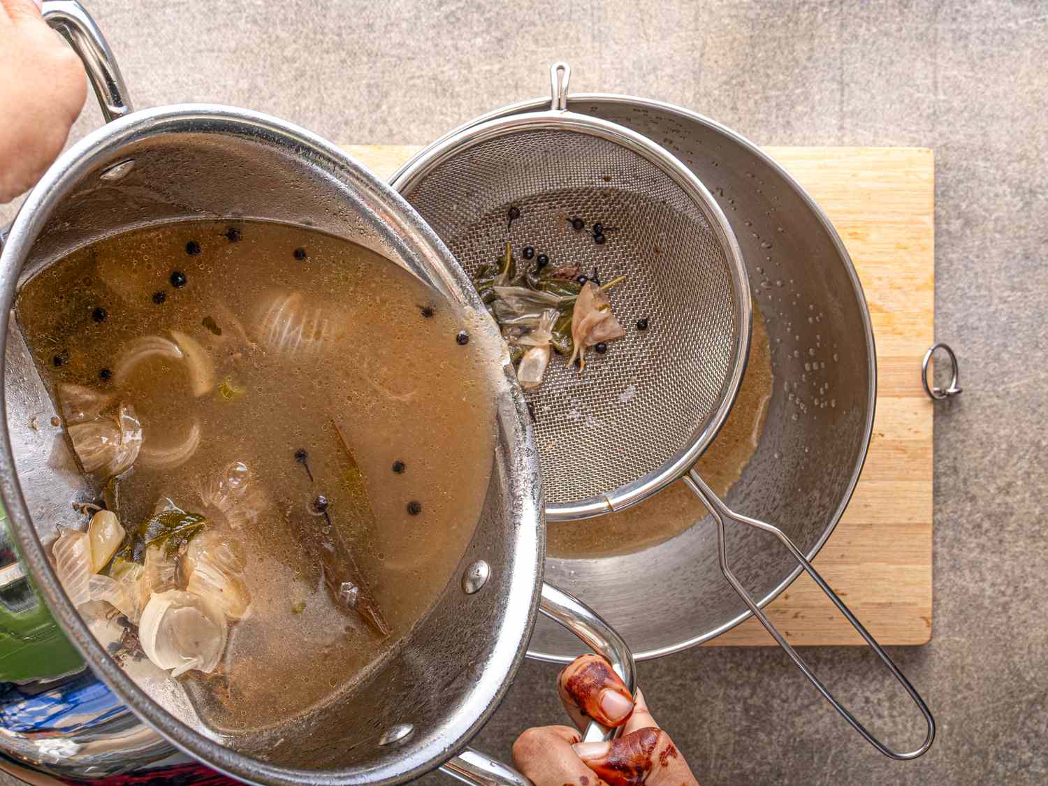 Straining broth through a sieve into a bowl with visible ingredients like peppercorns and herbs