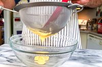 A person sifting a yellow liquid through the Rösle Stainless Steel Round Handle Kitchen Strainer into a glass bowl in a kitchen setting with an apron visible