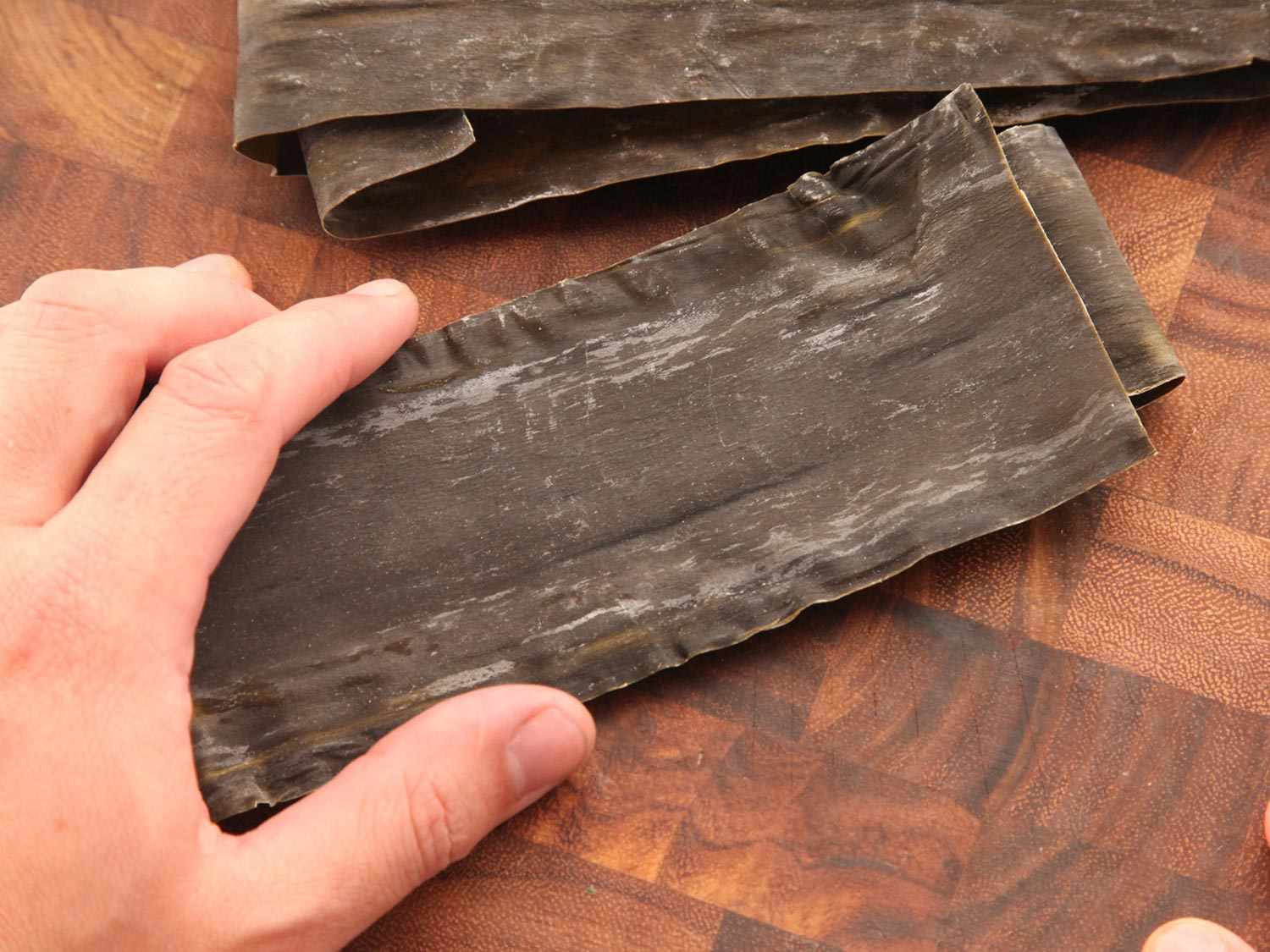 Hand holding strips of kombu on a wooden cutting board.