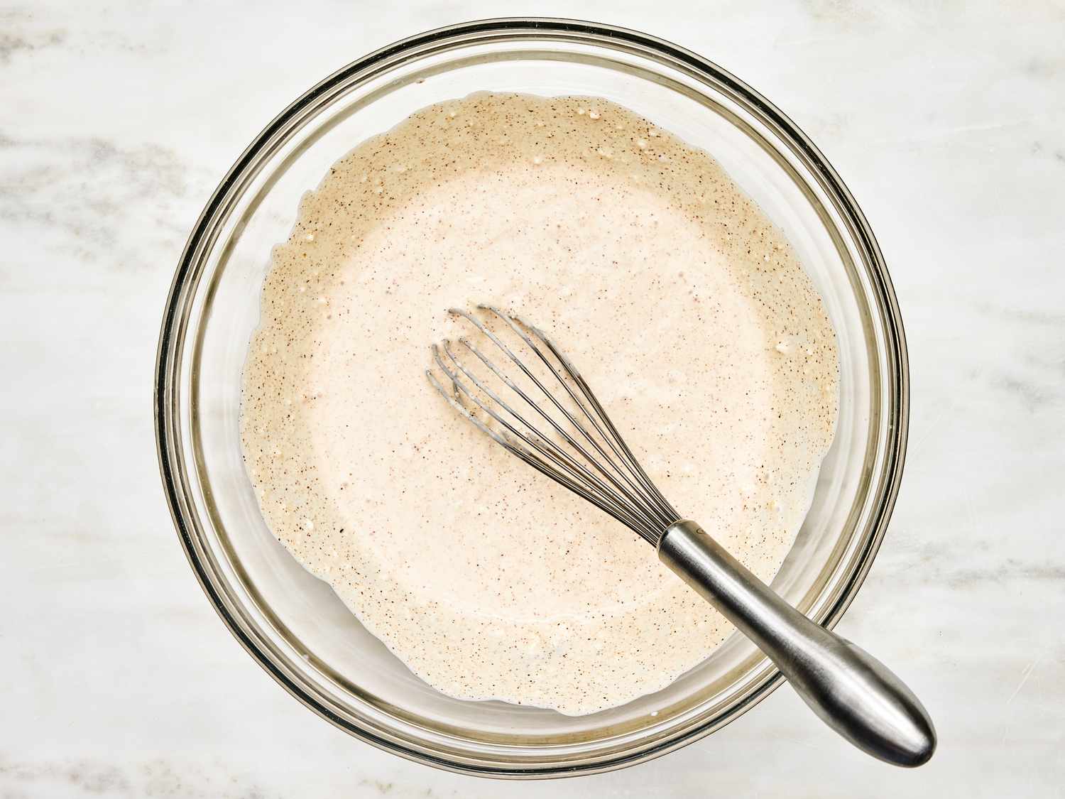 overhead angle of a clear glass bowl on a marble surface. Inside the bowl is a butter and cheese sauce wisked together with a wisk. 