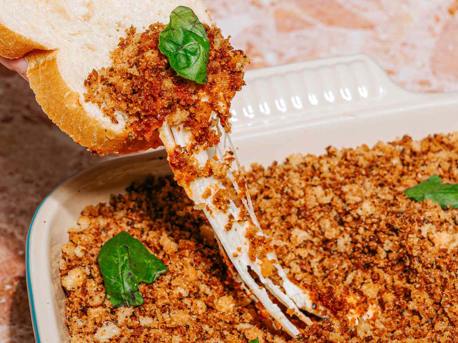 A bread scoop pulling melted mozzarella with breadcrumbs from a dish garnished with fresh basil