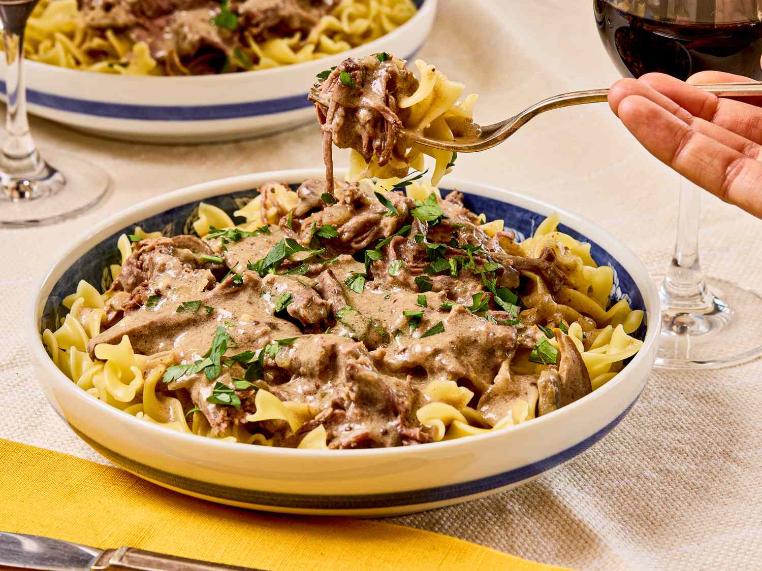 A fork lifting a portion of beef stroganoff served over egg noodles garnished with parsley