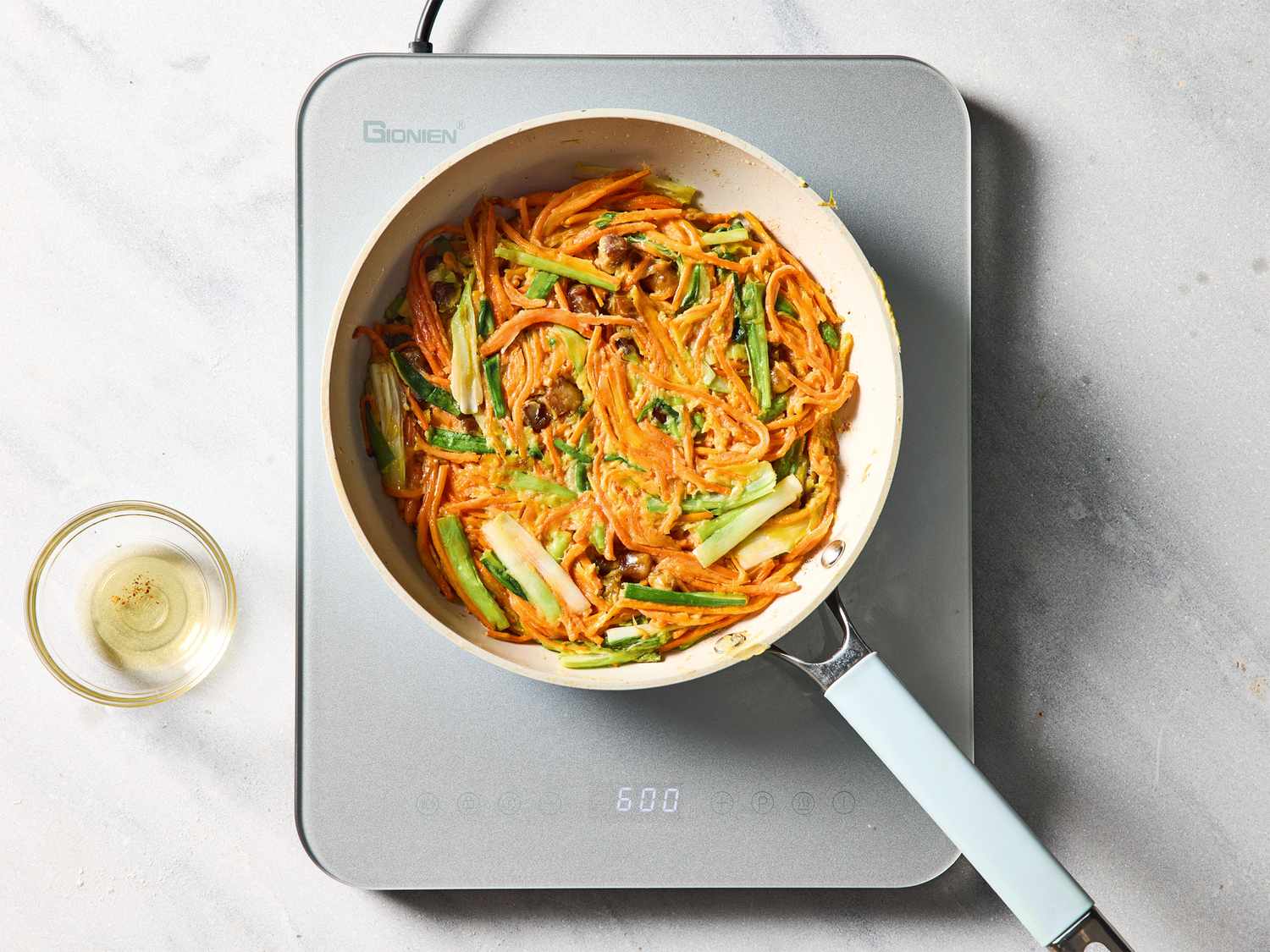 A skillet with carrots and greens cooking on a stovetop, a small glass bowl nearby
