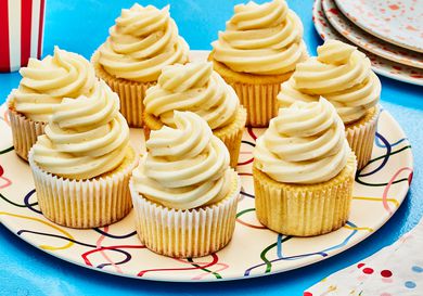 Horizontal image of 8 vanilla cupcakes on a colorful plate, and blue table. To the side is a stack of serving plates and colorful napkins