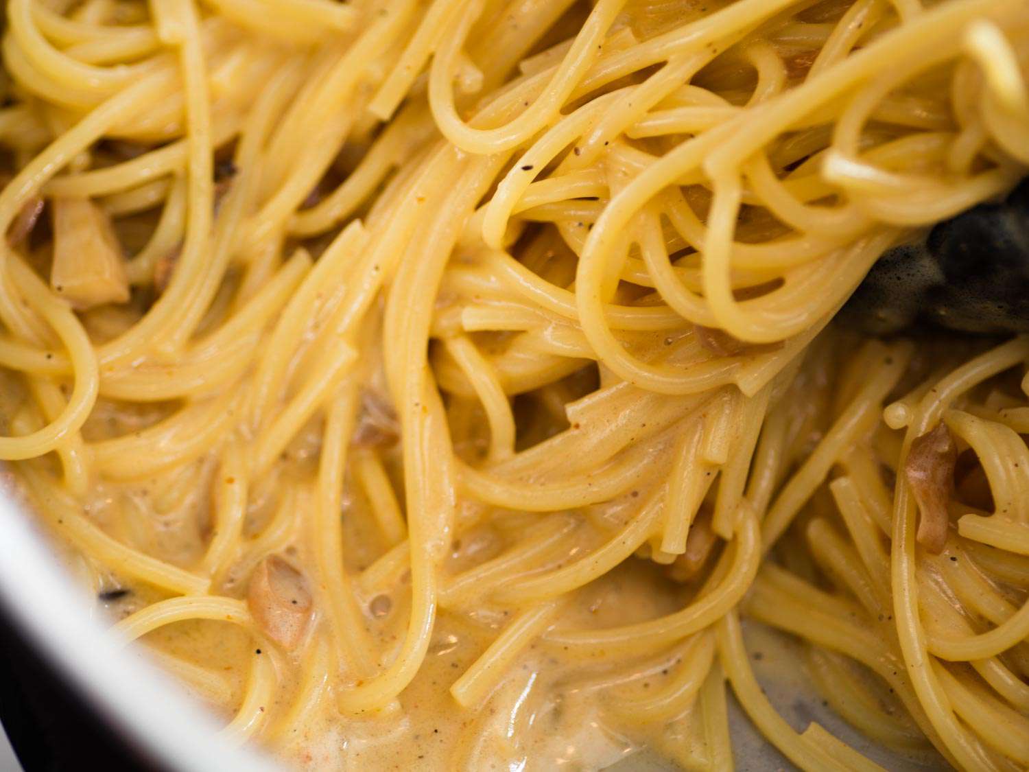Close-up of the simmering carbonara: par-cooked spaghetti is coated and half-submerged in a creamy, pepper-flecked sauce.
