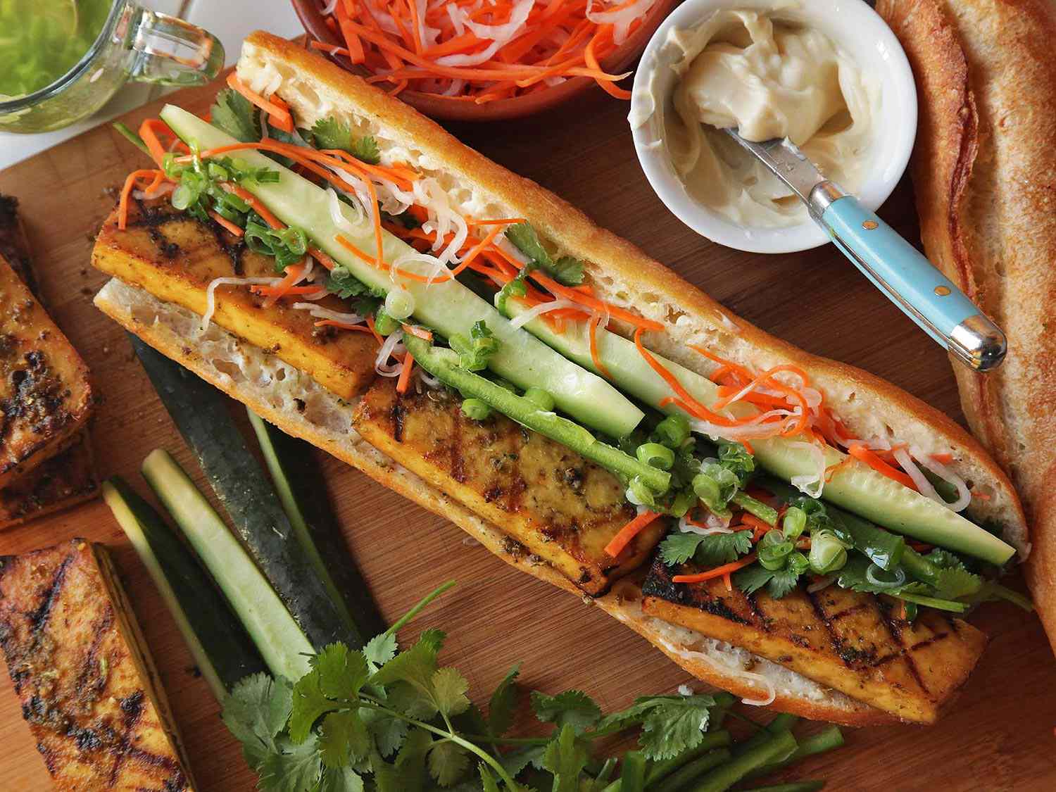 A Lemongrass-Marinated Tofu Banh Mi Sandwich on a cutting board, with extra toppings around it. 