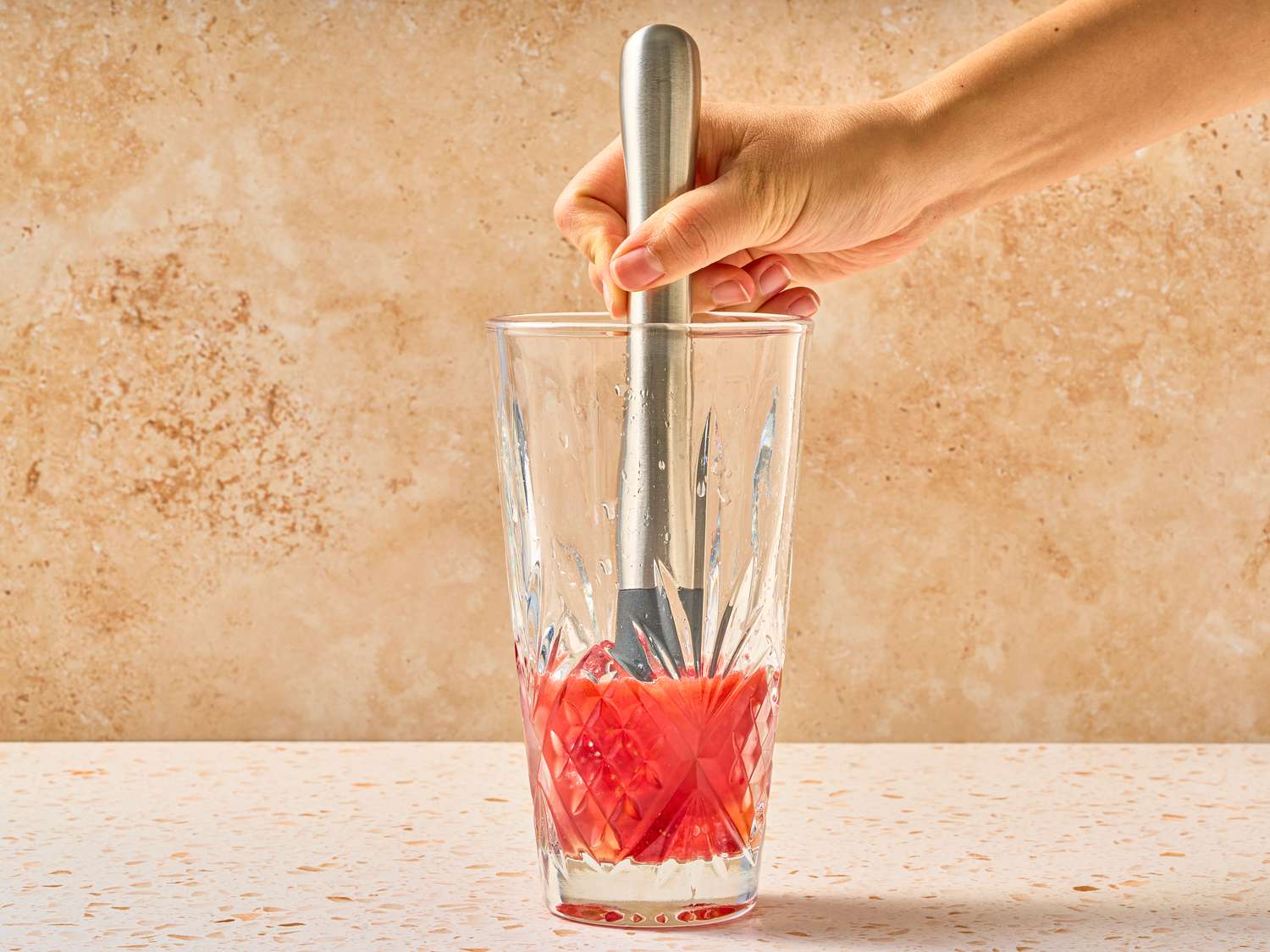 A hand using a muddler in a glass containing sliced strawberries on a countertop