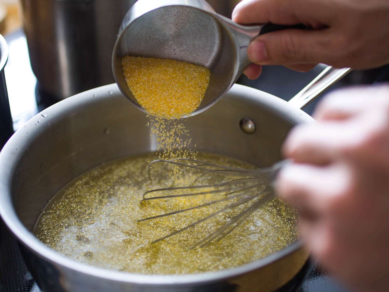 Pouring polenta from a measuring cup into a pot of water and whisking.