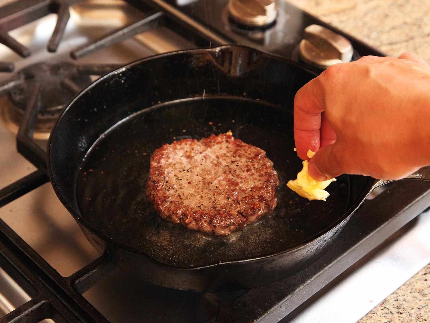 Adding butter to a cast iron skillet that contains a sous vide hamburger in the process of searing.
