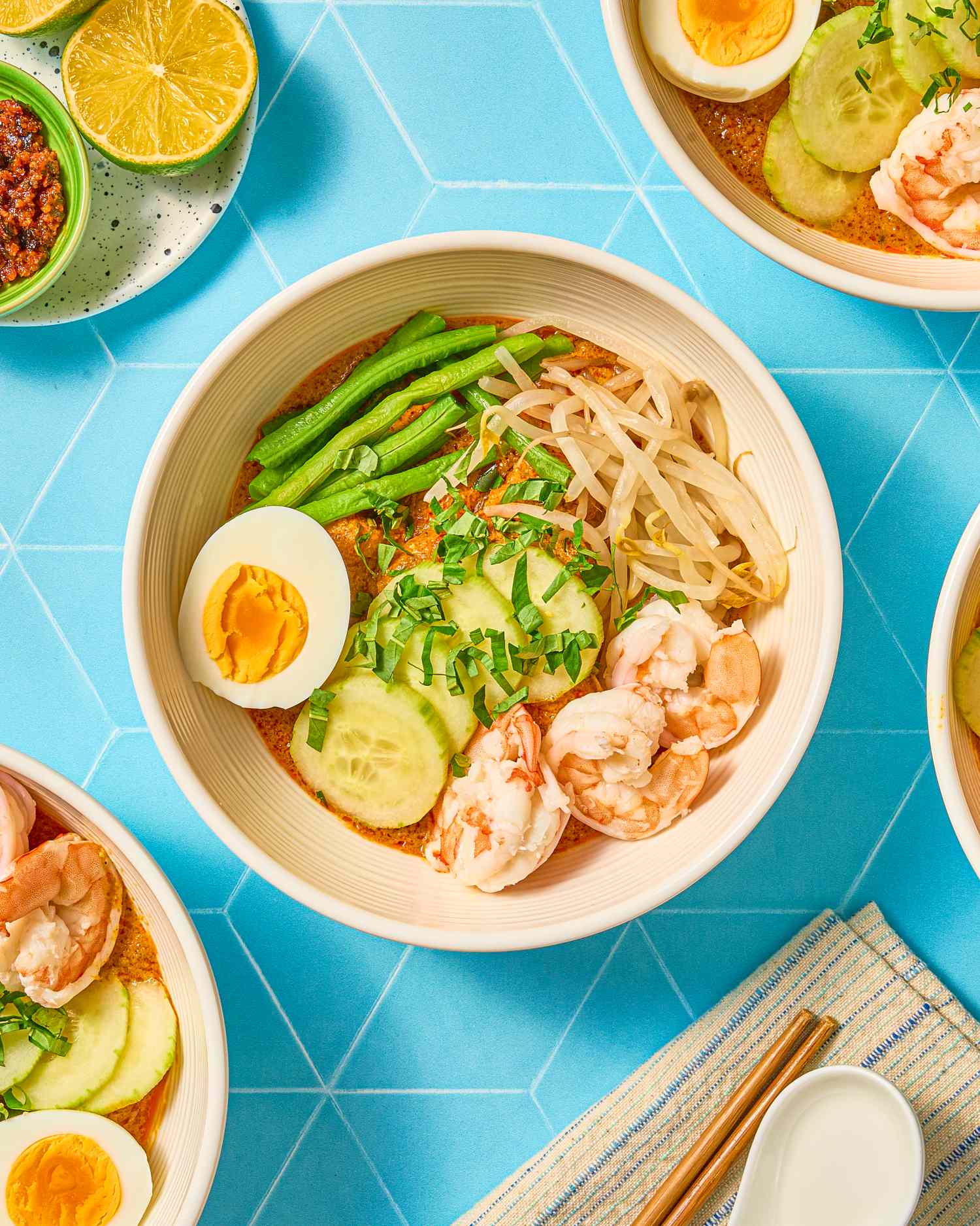 Bowl of curry laksa with toppings including shrimp egg and cucumber served on a blue geometric background