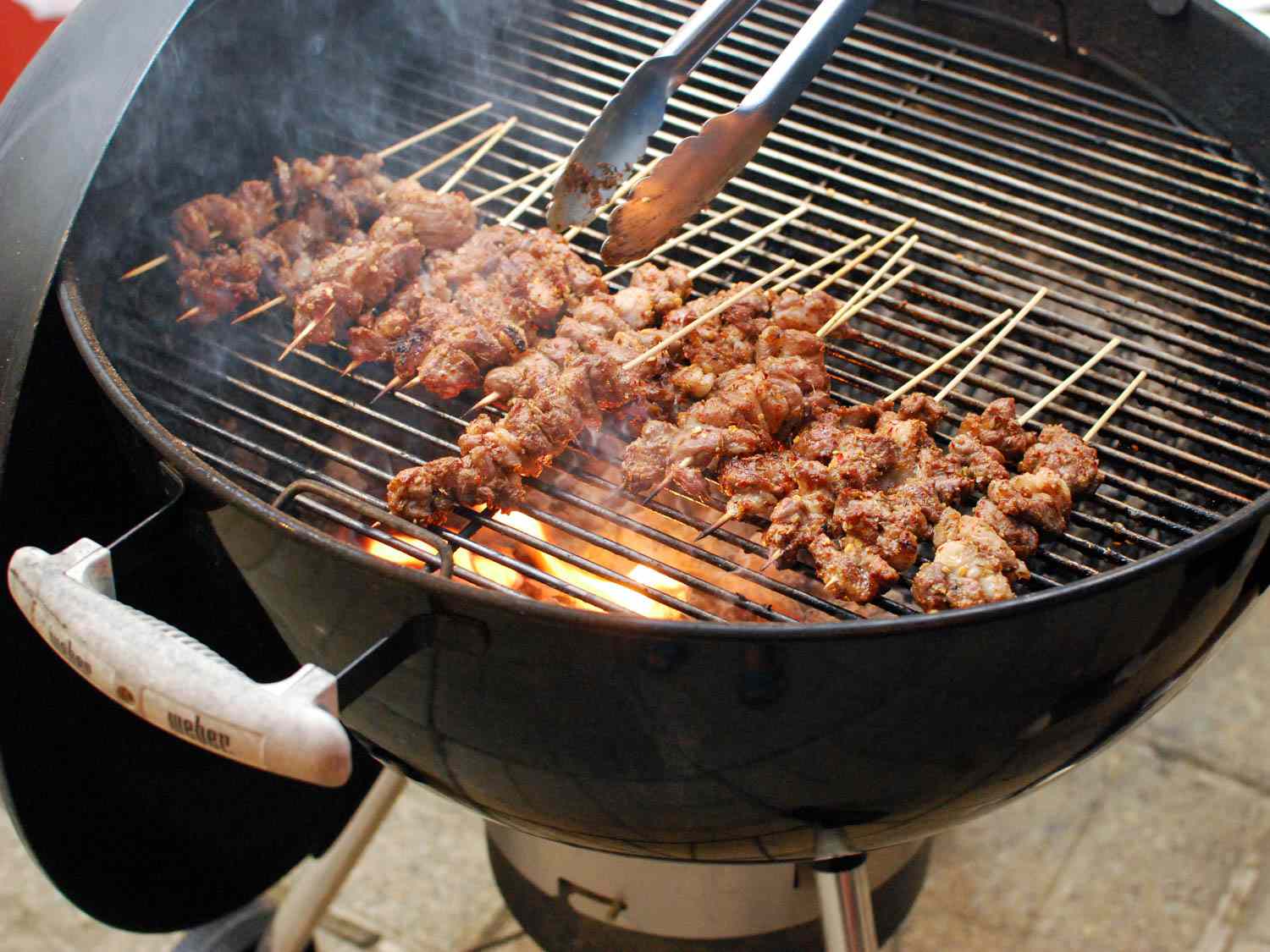 Using tongs to turn cooking lamb skewers on a grill.