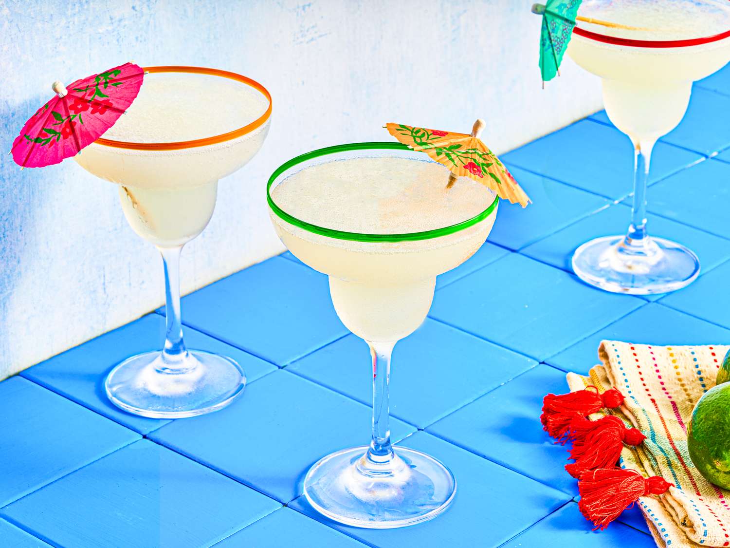 Three margaritas on a blue tile surface, each garnished with small paper umbrellas, with a decorated napkin and lime nearby