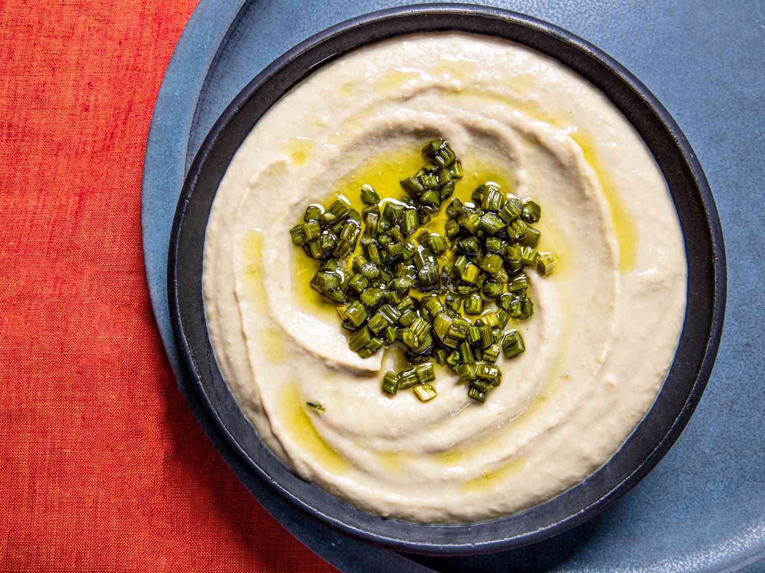 Garlic scape hummus topped with olive oil and garlic scape bits in a dark blue bowl placed off center of a light blue plate, on a red table cloth