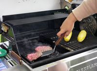 a person scraping up smash burgers from a griddle with corn on the grill too