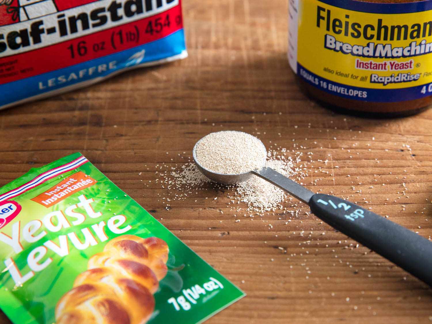 Three different kinds of yeast on a wooden surface with a half teaspoon full of yeast. 