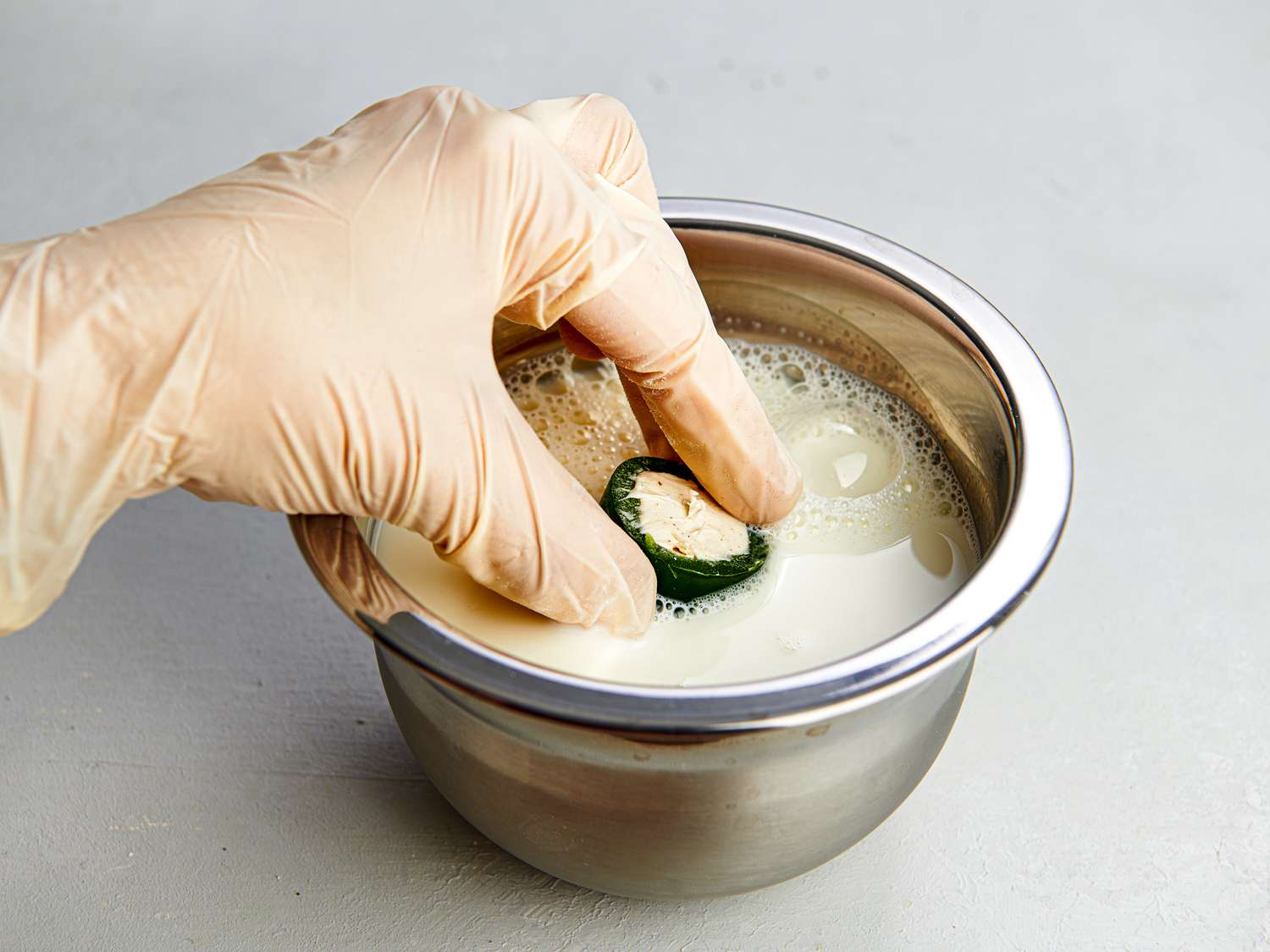 A gloved hand dipping a stuffed jalapeÃ±o ring into a small metal mixing bowl filled with milk.