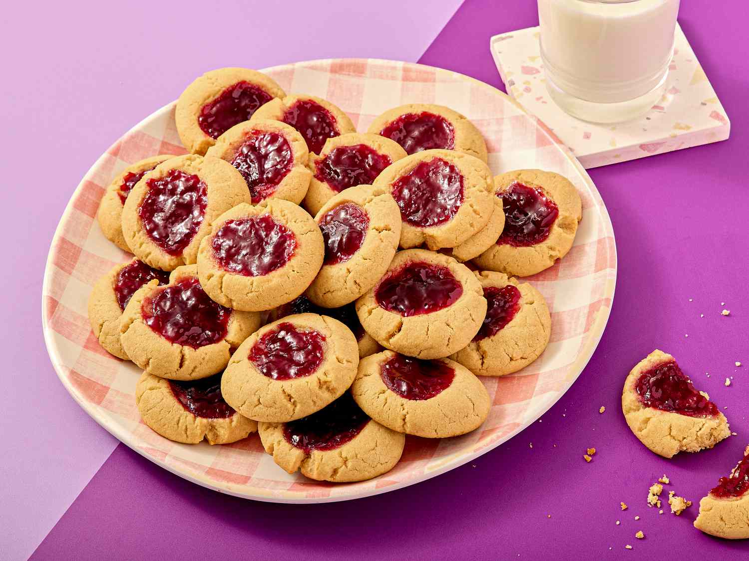 A plate of thumbprint cookies with jam filling next to a glass of milk and one partially eaten cookie