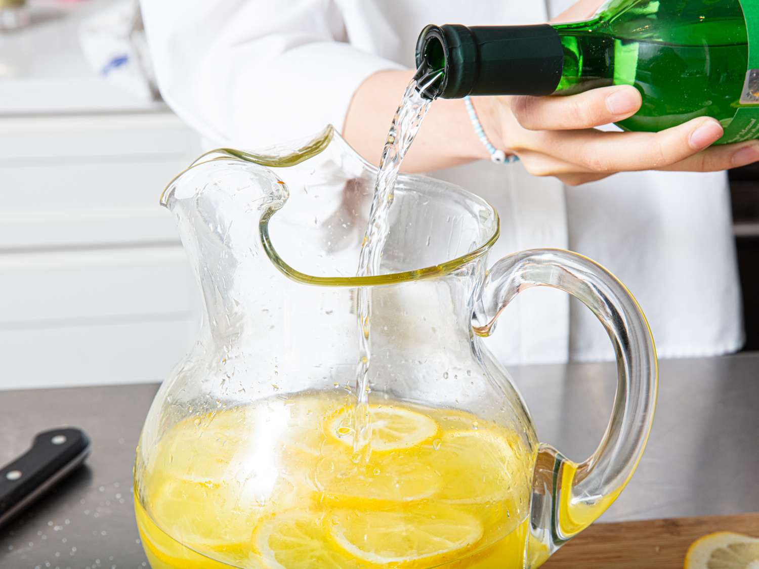 A pitcher filled with lemonade and lemon slices a person pouring a green bottles contents into it