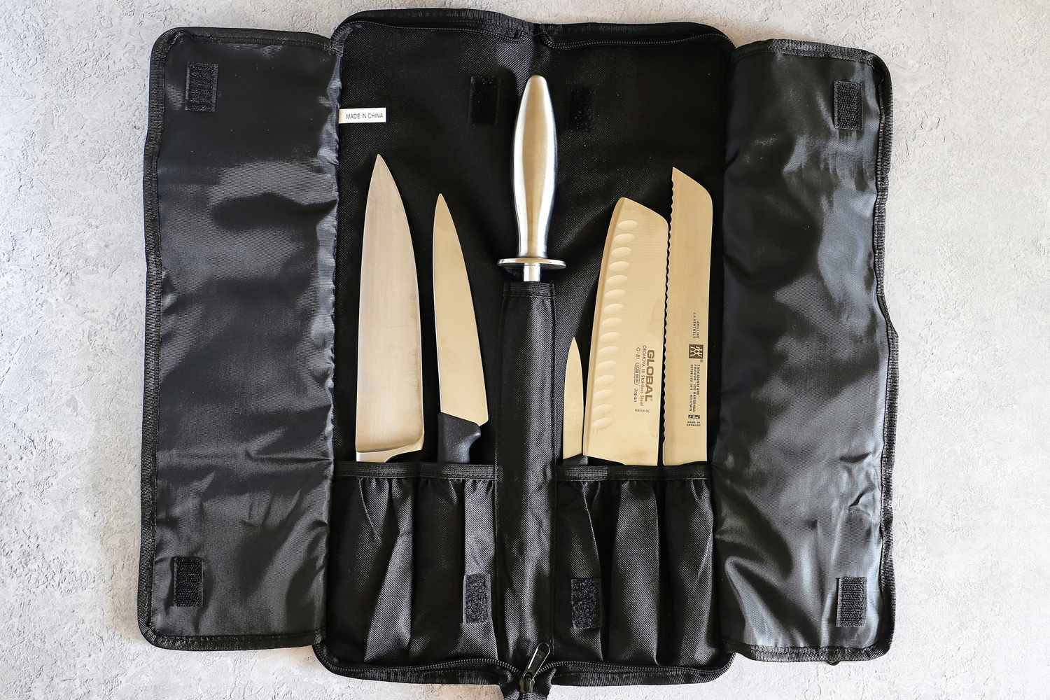 A set of kitchen knives and a sharpening steel organized in a black Asaya Chef Knife Roll Bag