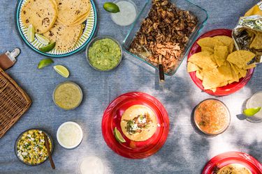 Snacks, dips, tortillas, lime wedges, salads, and dips as part of a Mexican-themed picnic spread on a picnic blanket. 