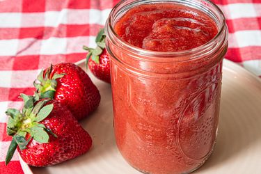 Glass jar of jam with whole strawberries on a plate and checkered tablecloth