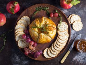 Overhead view of Baked Brie en Croûte With Thyme and Fig Jam, served on a wooden platter surrounded by grapes, shingled crackers, and herbs.