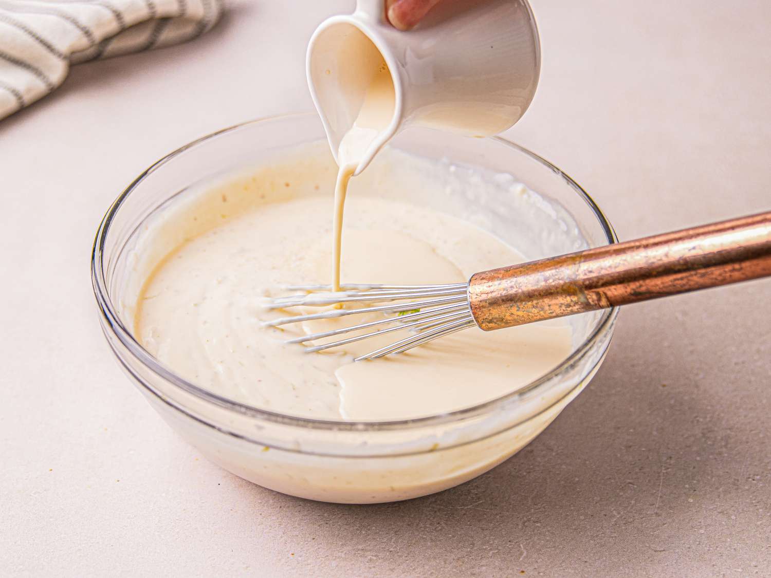 A mixing bowl with a whisk and creamy liquid being poured in from a small pitcher
